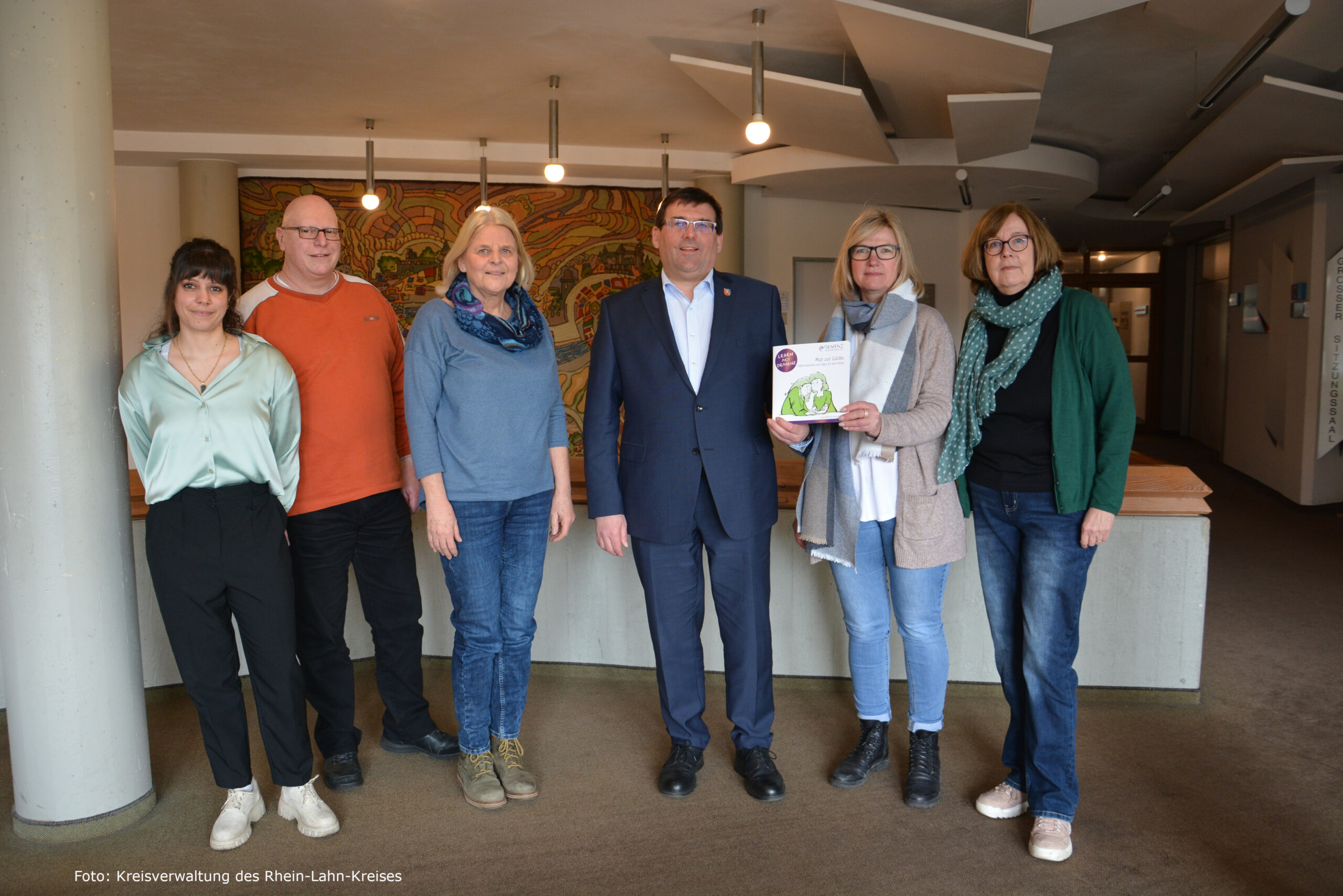 20230316 Broschürenvorstellung mit Quellenangabe Gruppenfoto von sechs Personen bei der Vorstellung der Broschüre ‚Leben mit Demenz‘ im Gebäude der Kreisverwaltung Rhein-Lahn. Eine Frau hält die Broschüre in der Hand.
